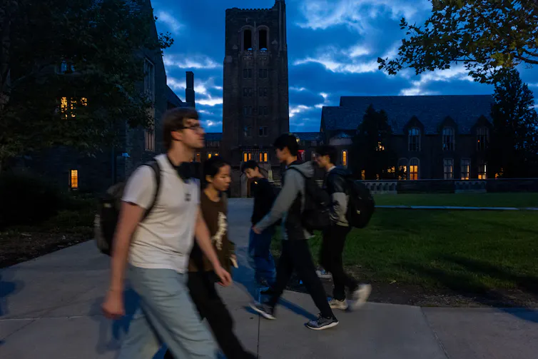 Groups of young people walk in front of buildings at dusk, with a dark blue sky.