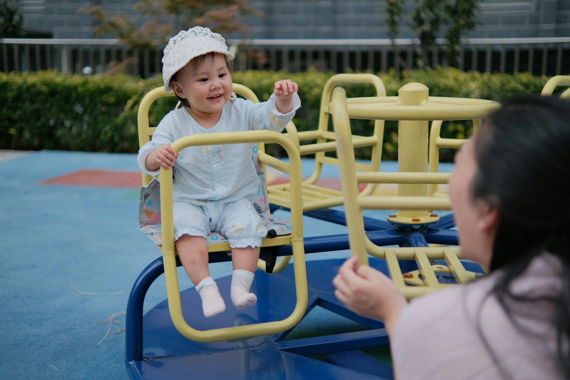 A preschooler on a play structure waving at an adult who is responding with raised hand.