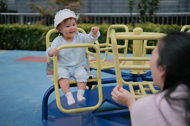 A preschooler on a play structure waving at an adult who is responding with raised hand.