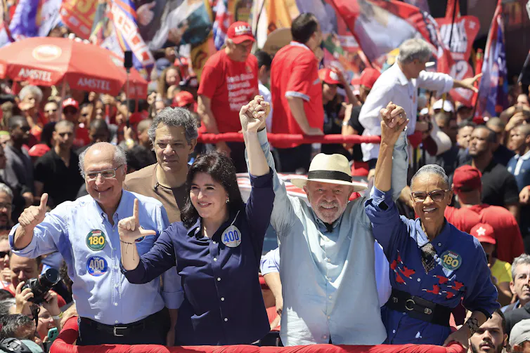 Brazilian President Luiz Inácio Lula da Silva smiles and holds hands with senior political figures on a float, surrounded by supporters waving flags at a campaign rally.