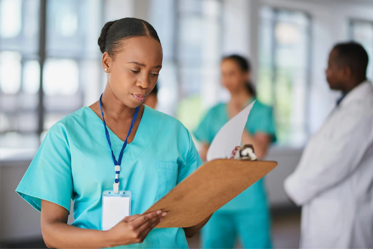 (A Black doctor in scrubs with a stethoscope looking at a clipboard with other health-care professionals out of focus in background)