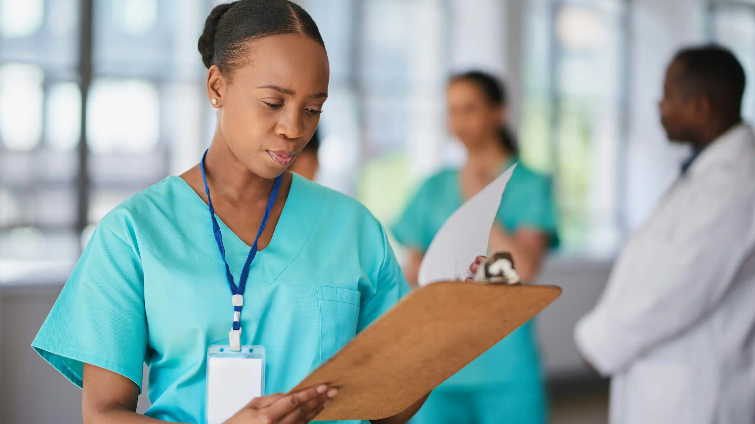 (A Black doctor in scrubs with a stethoscope looking at a clipboard with other health-care professionals out of focus in background)