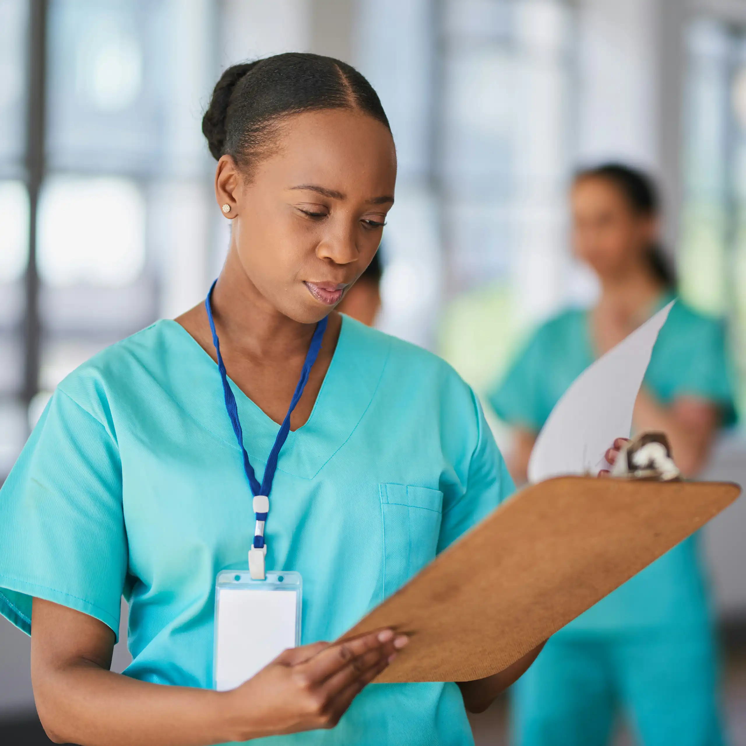 (A Black doctor in scrubs with a stethoscope looking at a clipboard with other health-care professionals out of focus in background)