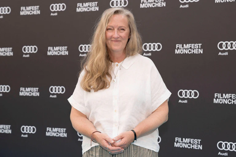 A middle-aged woman with long blonde hair stands smiling at the camera.