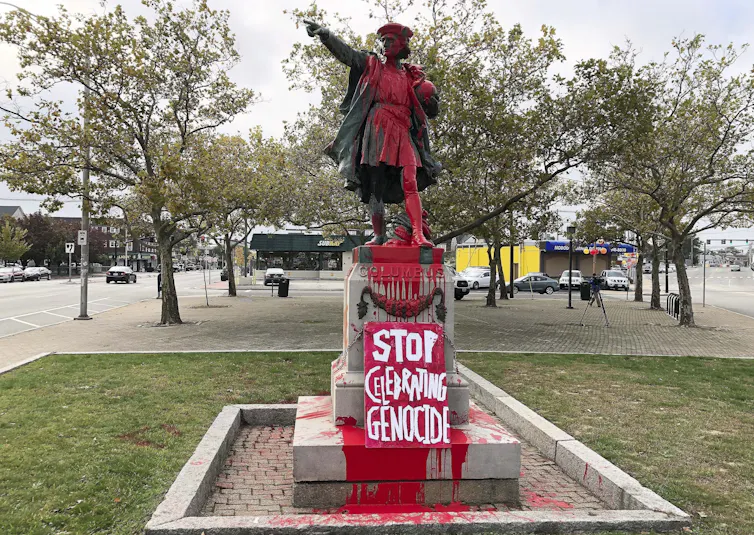 A statue of a man with an outstretched arm is covered in red paint and the words 'stop celebrating genocide.'