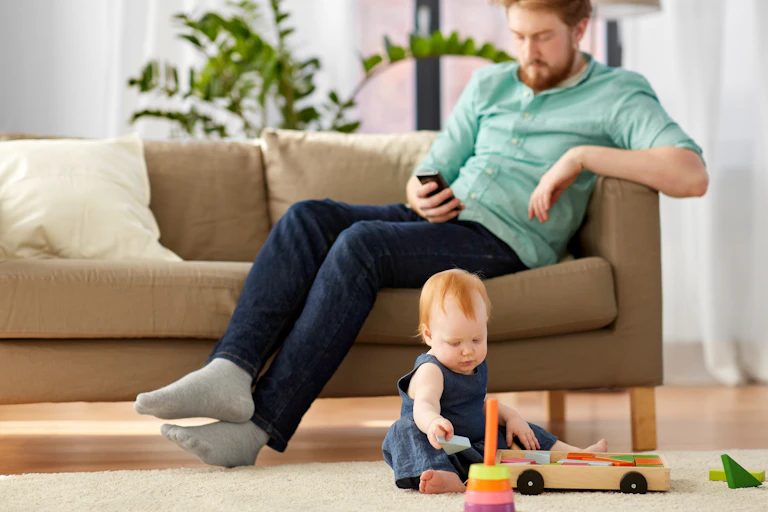 A father on his cellphone while a child plays with a toys on a carpet.