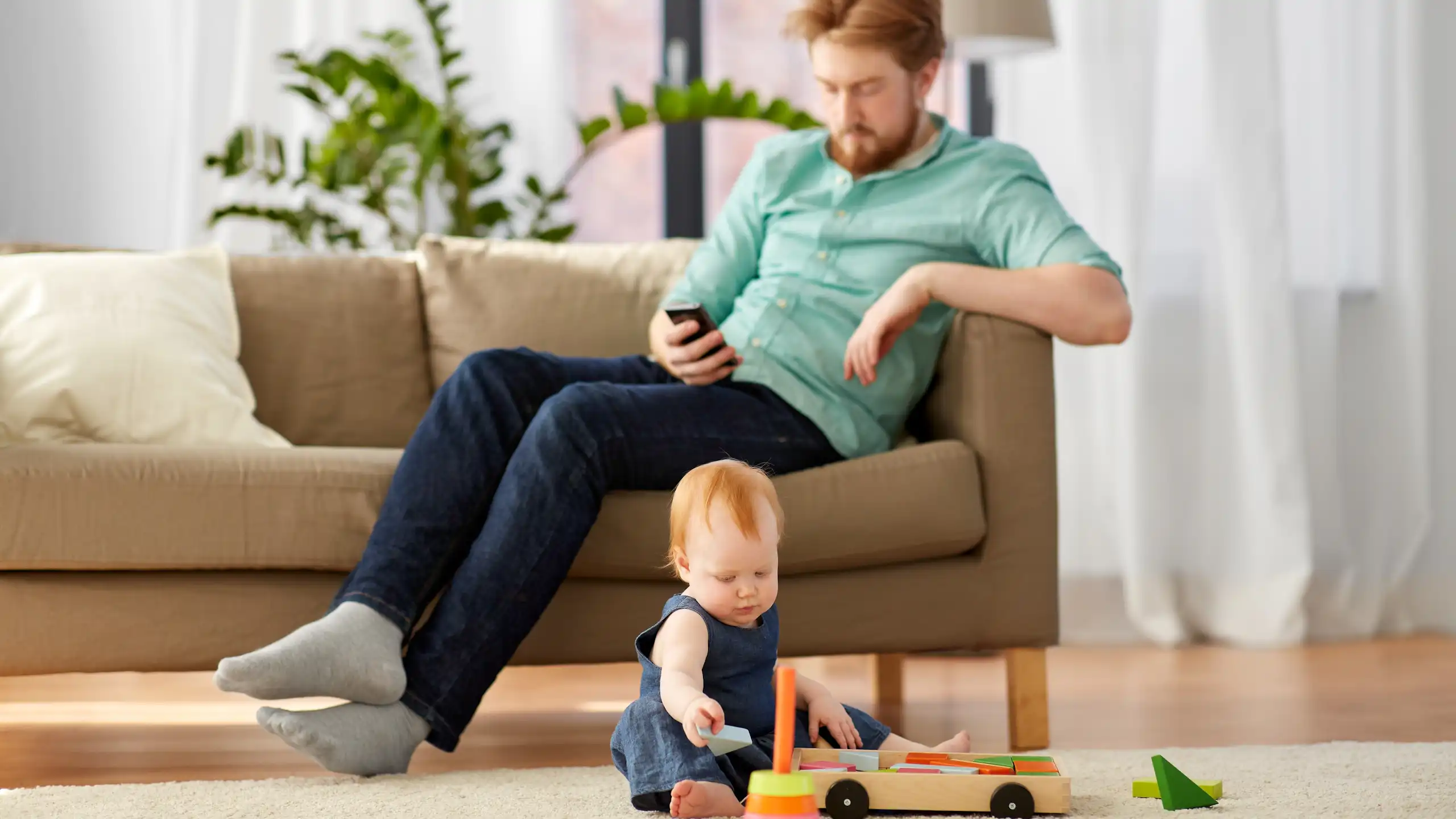 A father on his cellphone while a child plays with a toys on a carpet.
