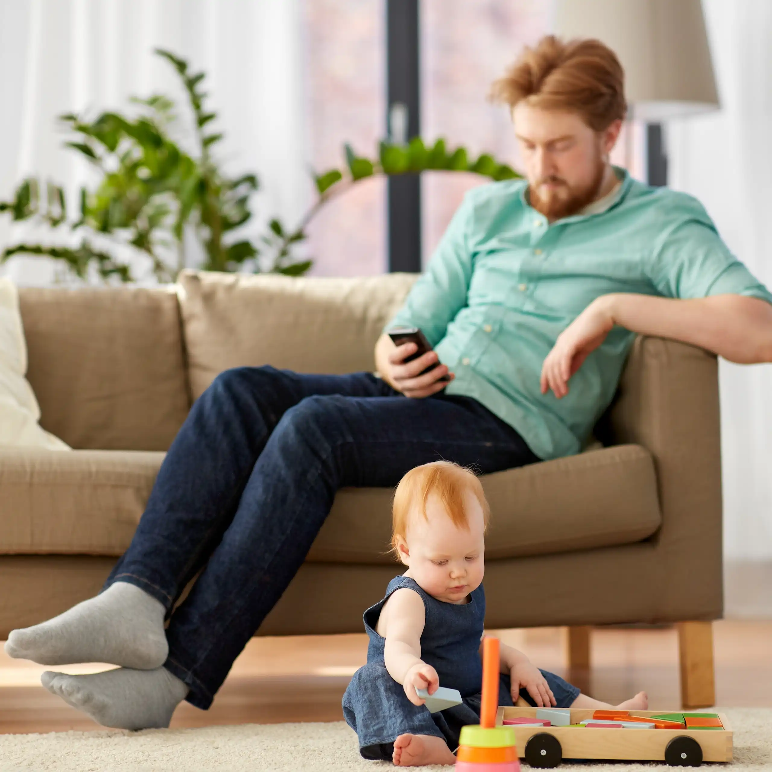 A father on his cellphone while a child plays with a toys on a carpet.
