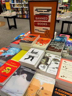 A table with stacks of colorful books surrounding a sign reading 'Banned Books.'