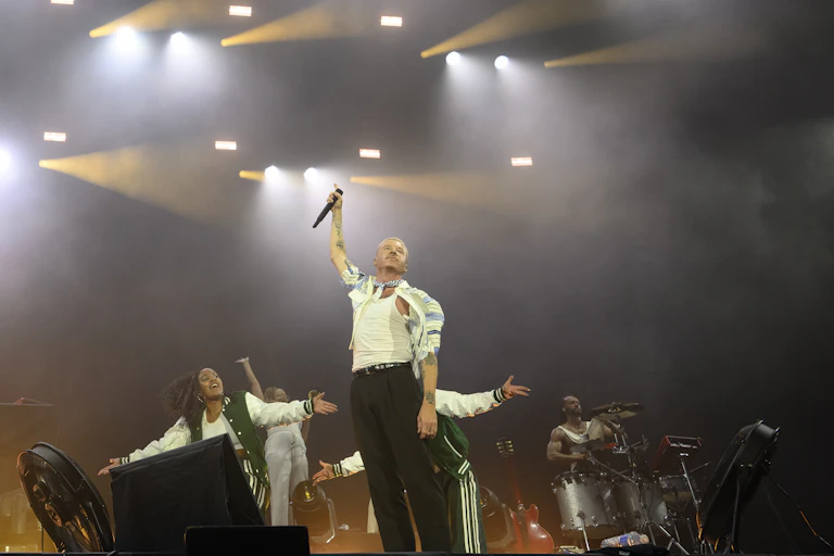 A young man raises a microphone into the air from the stage, surrounded by bandmates.