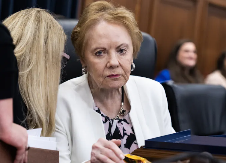 An older woman in a white blazer stares somewhat blankly as a younger woman briefs her.