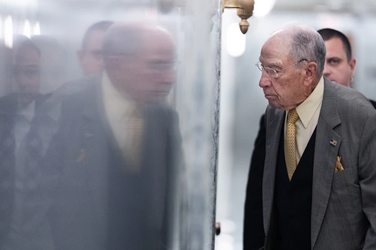 An older man in blazer and vest stands before a shiny surface that reflects his image.