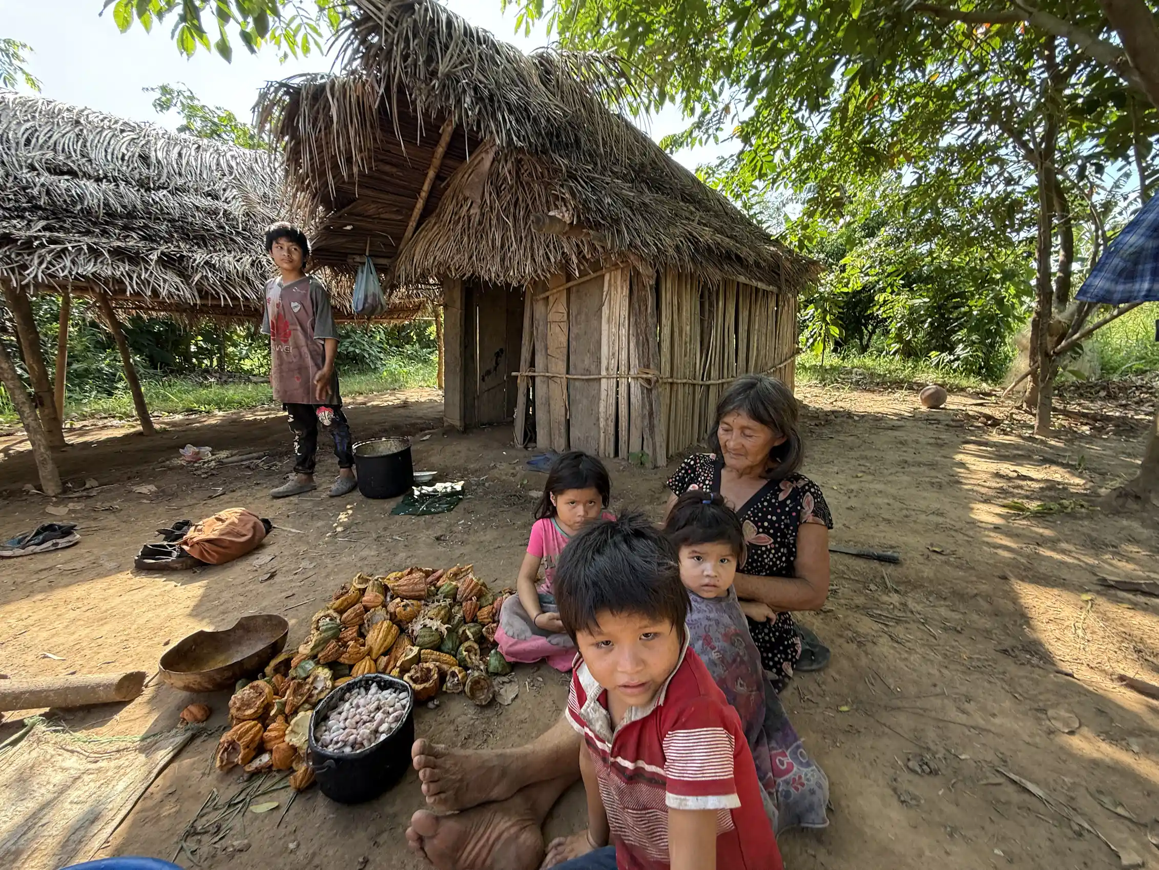 several children sit with a woman next to some harvested crops in front of a thatched hut