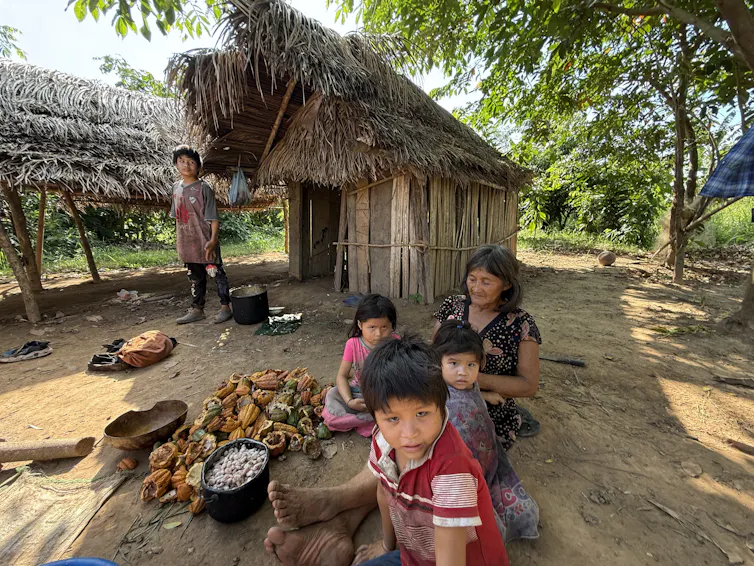 several children sit with a woman next to some harvested crops in front of a thatched hut