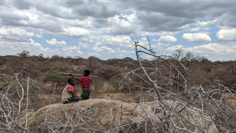 Two Hadza children play atop a rocky outcrop, with bushland and clouds in the background