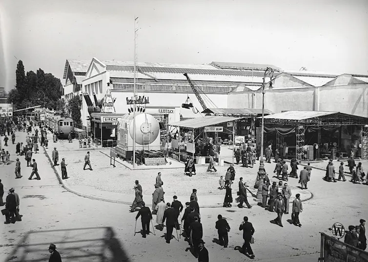 historic photo of people at an outdoors trade fair with large electric equipment at centre.
