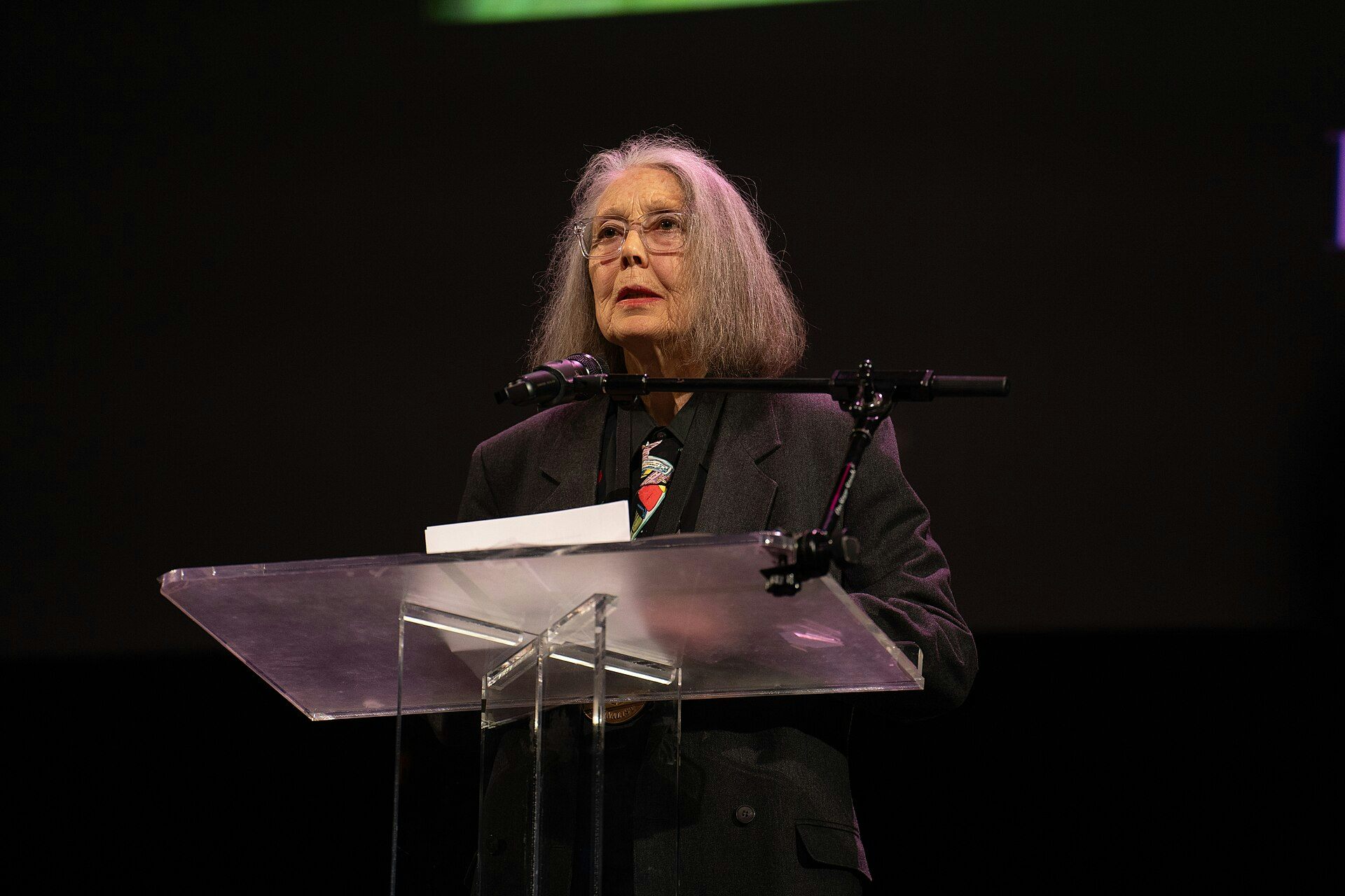 A woman at a podium reads against a dark background.