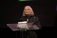 A woman at a podium reads against a dark background.