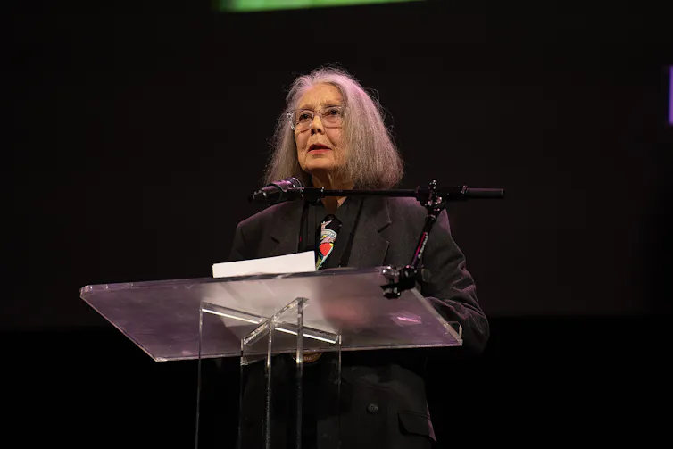 A woman at a podium reads against a dark background.