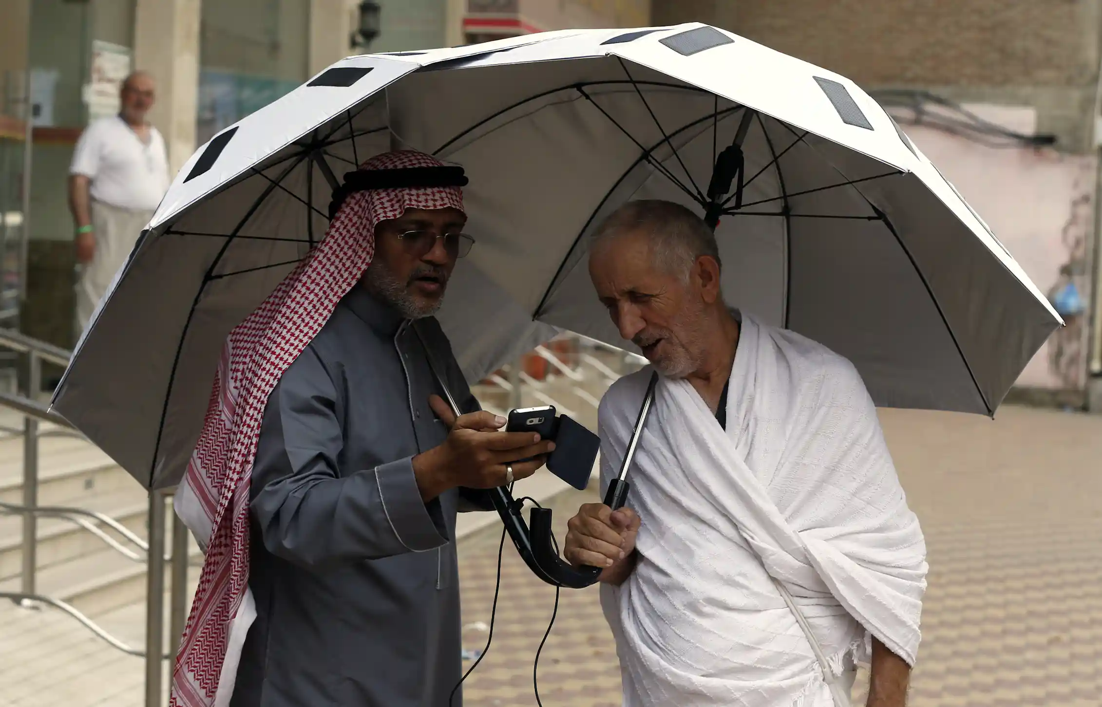 Two men stand under an umbrella as one shows the other something on a smartphone.