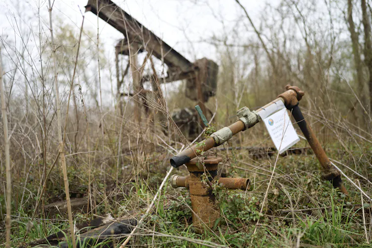 A rusting old piece of equipment sits in an area with low brush and a pipe sticking up out of the ground.