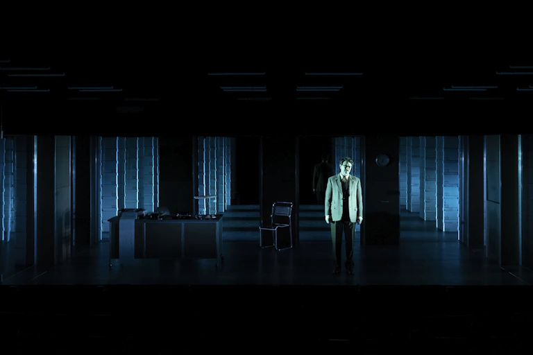 A man in a suit stands on a darkened stage with a grid of computer banks stretching out across the stage behind him.