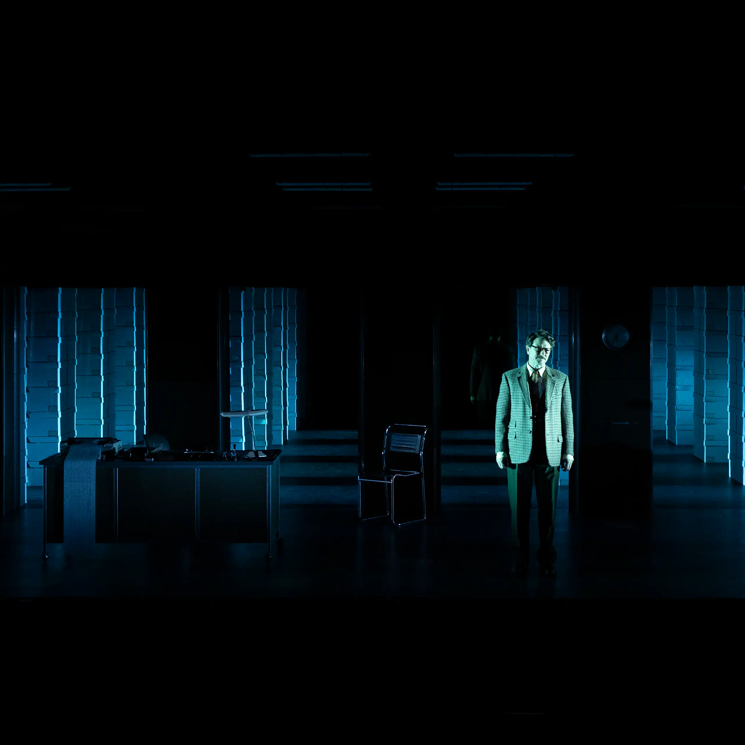 A man in a suit stands on a darkened stage with a grid of computer banks stretching out across the stage behind him.