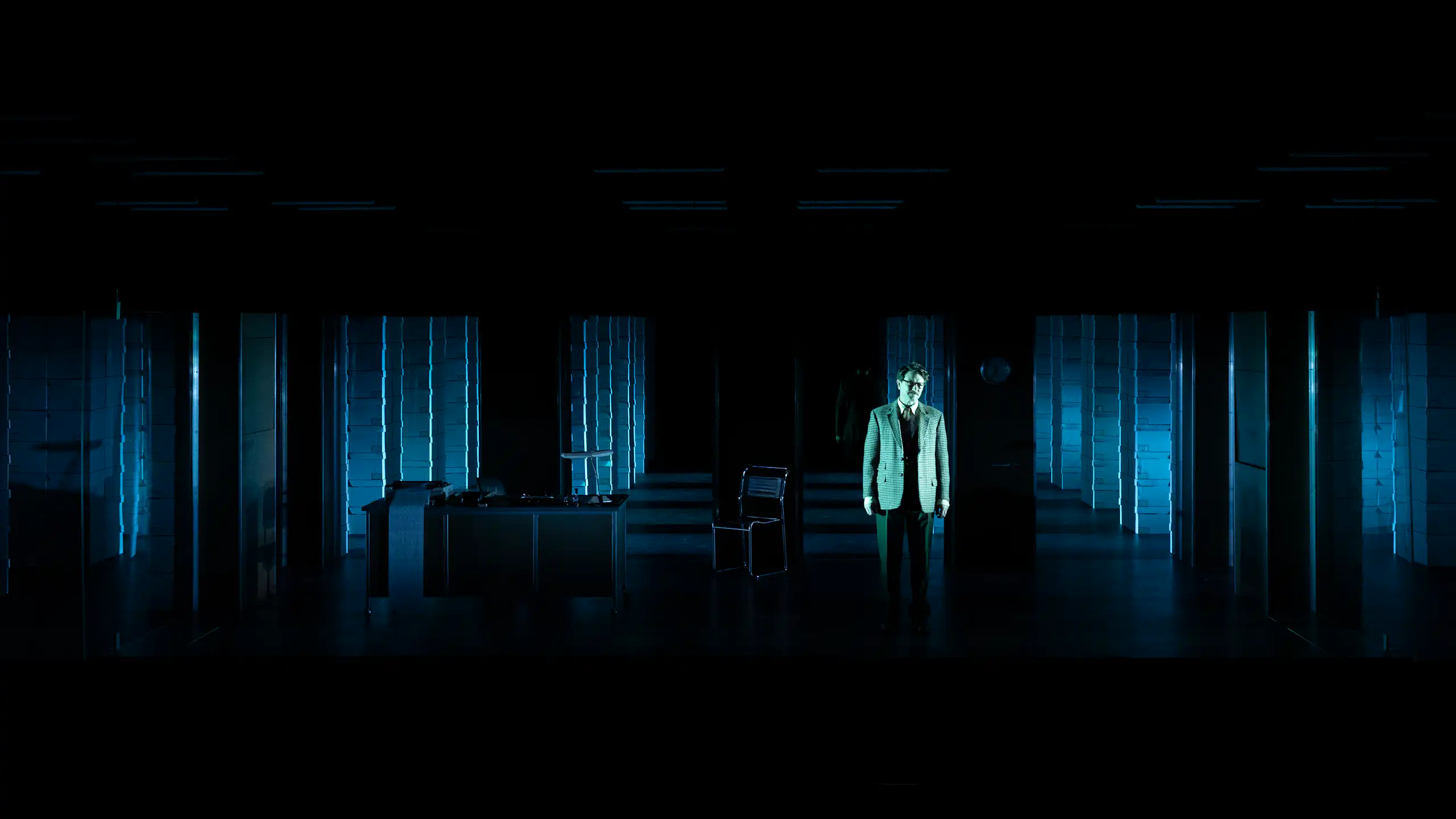 A man in a suit stands on a darkened stage with a grid of computer banks stretching out across the stage behind him.