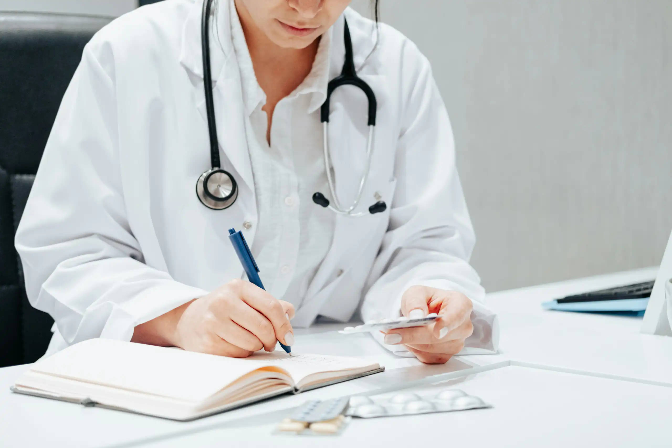 A person in a white coat and stethoscope writing, with a blister pack of medication in their other hand