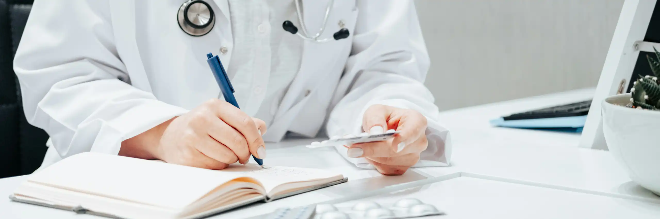 A person in a white coat and stethoscope writing, with a blister pack of medication in their other hand
