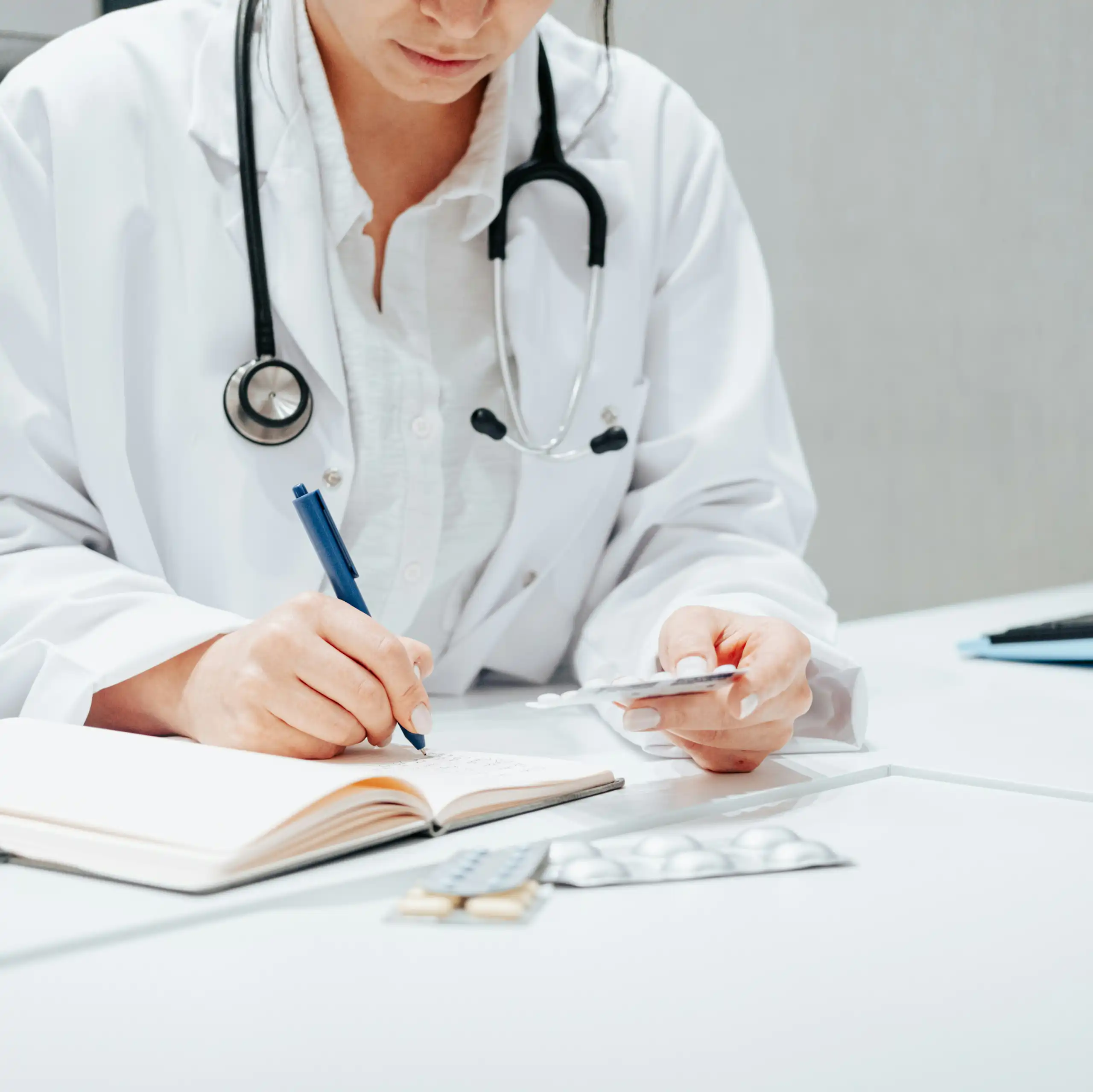 A person in a white coat and stethoscope writing, with a blister pack of medication in their other hand