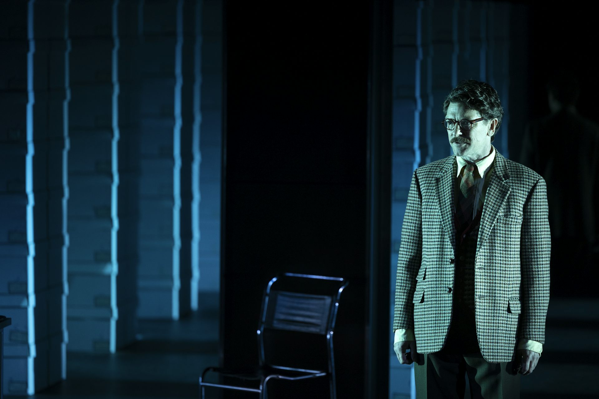 A man in a suit stands on a darkened stage with a grid of stacked document boxes behind him.
