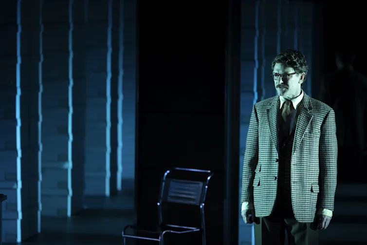 A man in a suit stands on a darkened stage with a grid of stacked document boxes behind him.