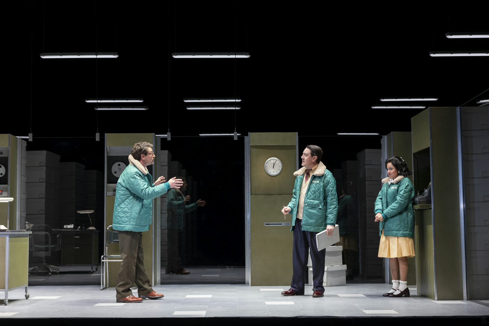 Three people in mint-coloured quilted jackets stand in a windowless office-like space with fluorescent lights and stacks of document boxes.
