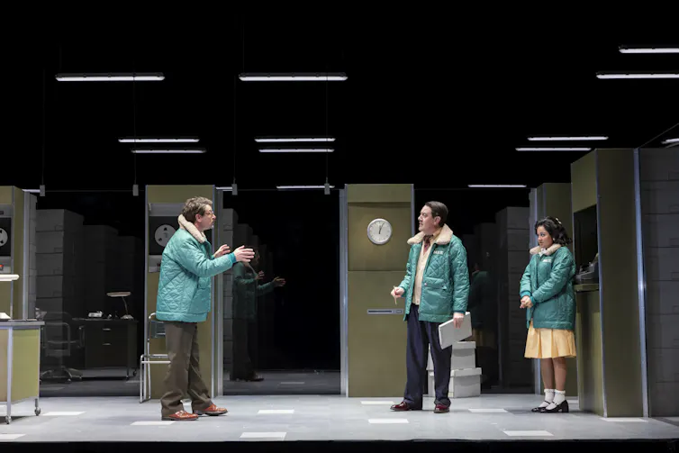 Three people in mint-coloured quilted jackets stand in a windowless office-like space with fluorescent lights and stacks of document boxes.