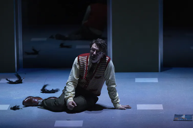 A man in a shirt and woollen vest slumps on stage with parts of black birds strewn around him.