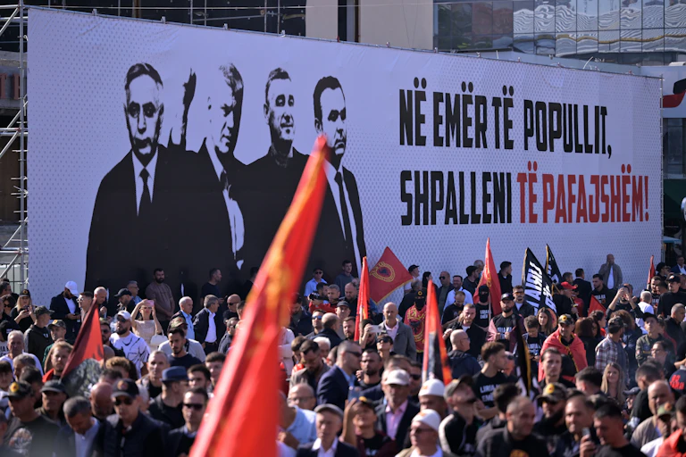 Protesers wave Kosovan flags in front of a billboard protesting the innocence of Hashim Thaci, Kadri Veseli, Rexhep Selimi, and Jakup Krasniqi