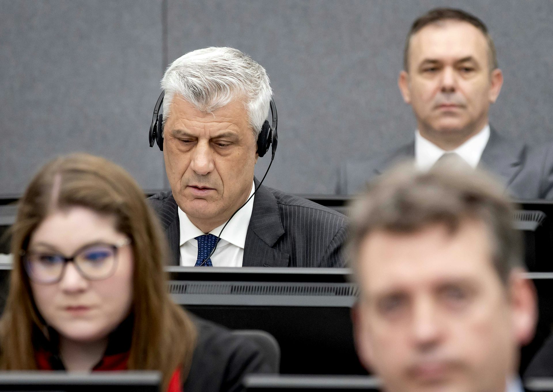 An elderly man sits in court wearing headphones. Behind him is another man in suit and a man and a woman site in front of him.