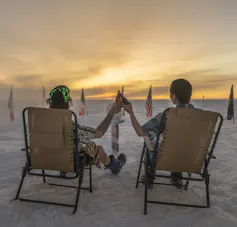 Two people sit in beach chairs on snow clinking bottles with the sun coming over horizon.