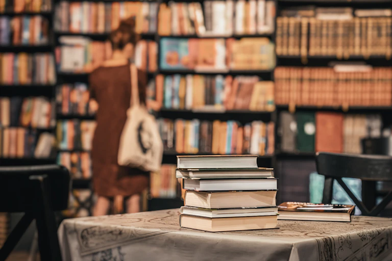 Quelques livres sur une table au premier plan, dans une librairie ou une bibliothèque où l'on voit une femme près des rayonnages.
