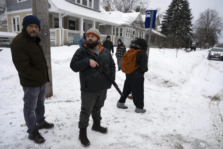 A man wearing a hat and coat holds an assault weapon as he stands on snow-covered ground.