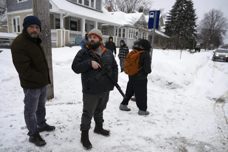 A man wearing a hat and coat holds an assault weapon as he stands on snow-covered ground.