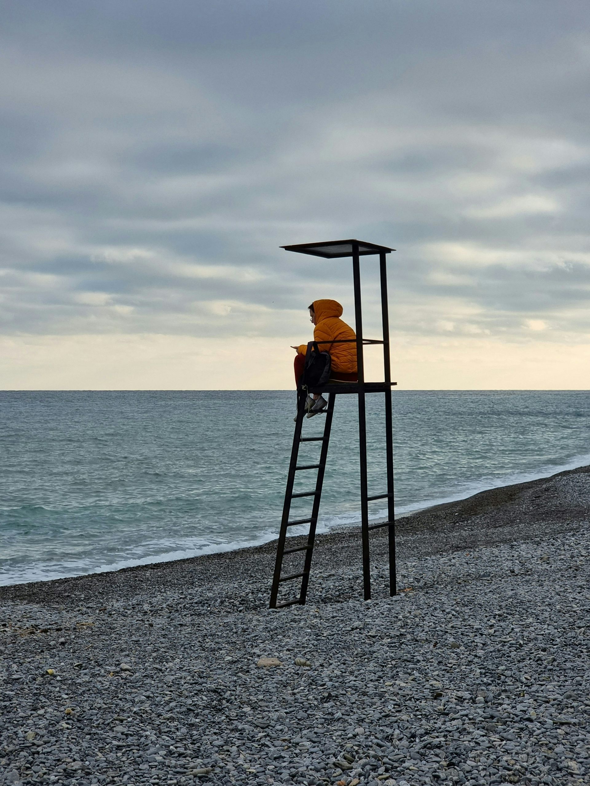 A lone lifeguard with an orange windbreaker sits atop his station, looking at the sea under an overcast sky.