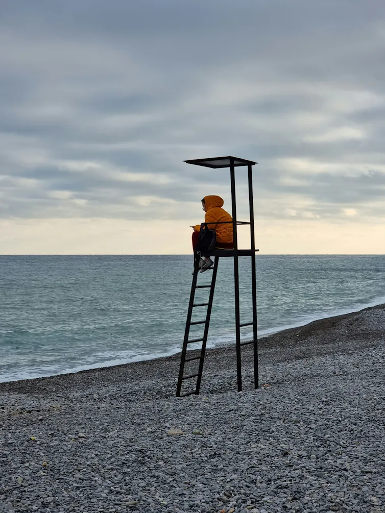 A lone lifeguard with an orange windbreaker sits atop his station, looking at the sea under an overcast sky.