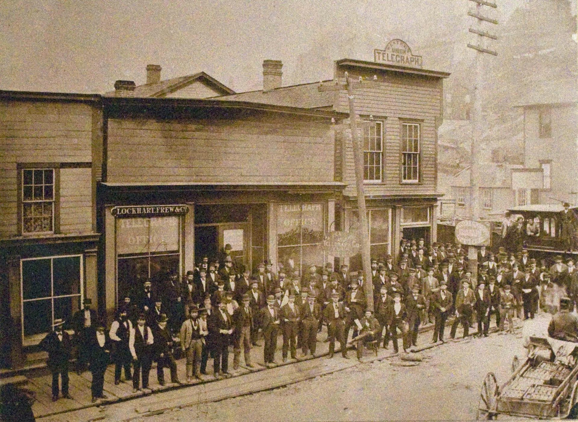 Dozens of men in suits stand along a curb in a sepia-toned photo.