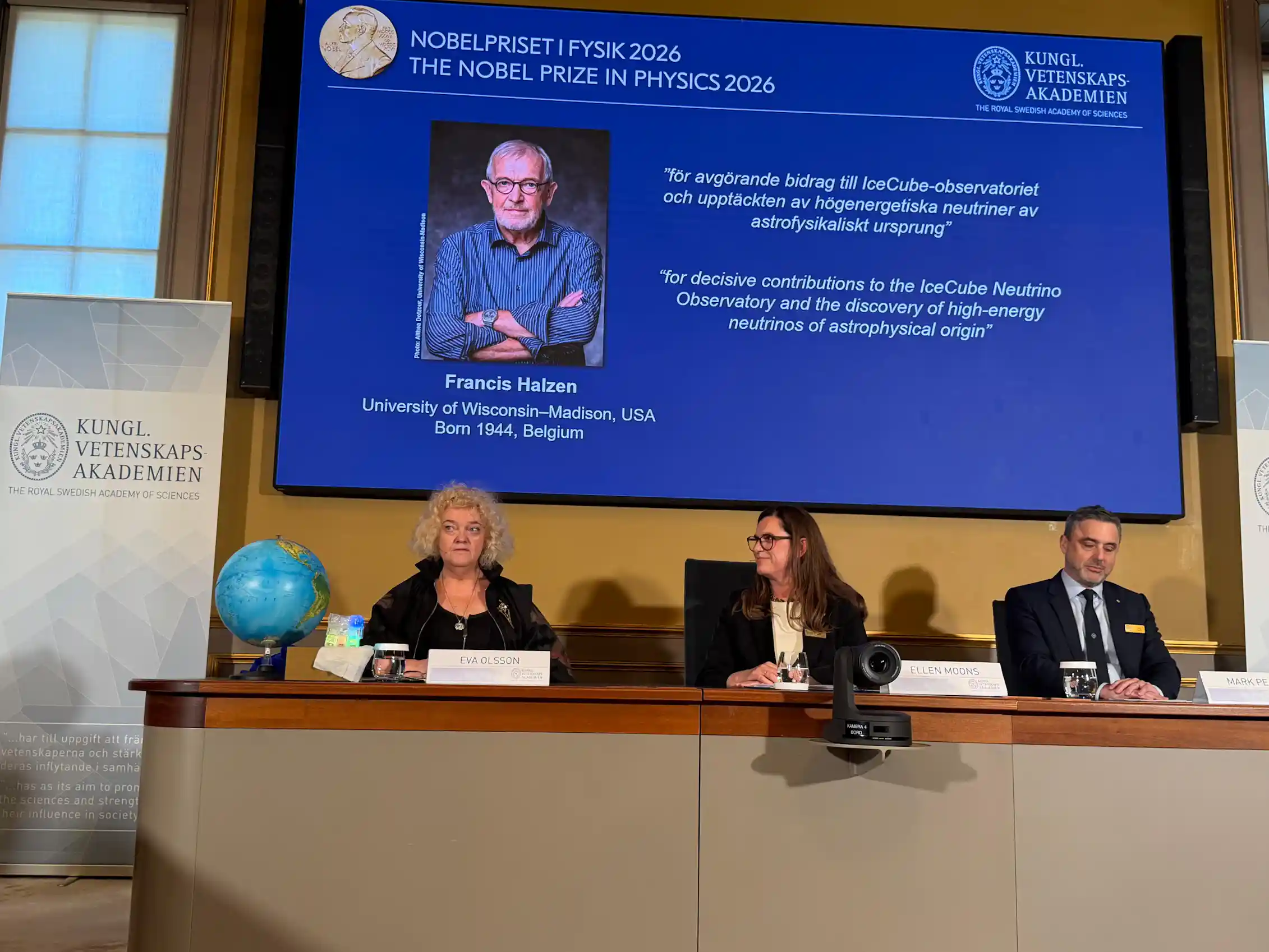 A panel of three people sitting at a table, with a screen behind them showing a photo of Francis Halzen and a description of his research.