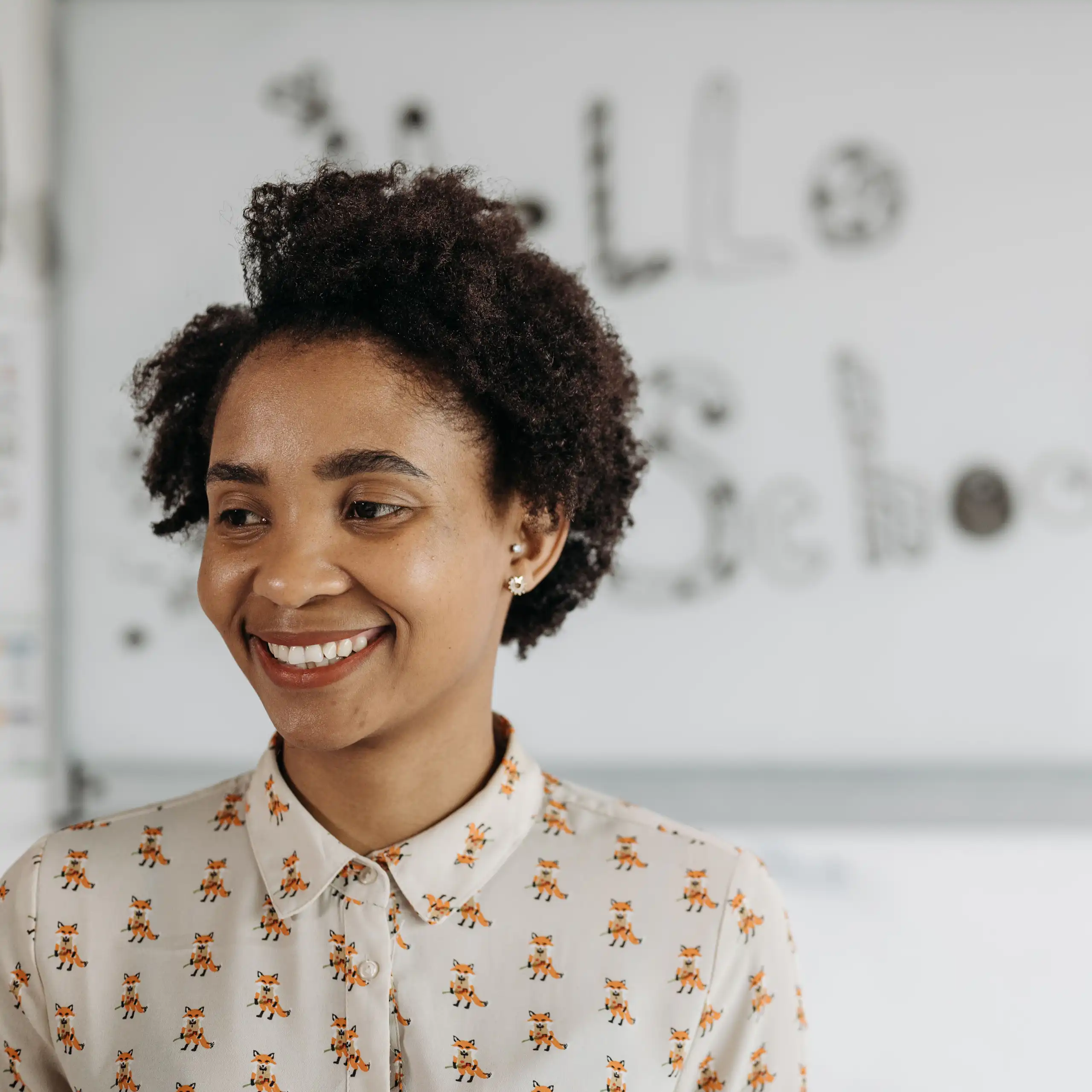 A woman stands in front of a whiteboard, smiling and looking off to the side.