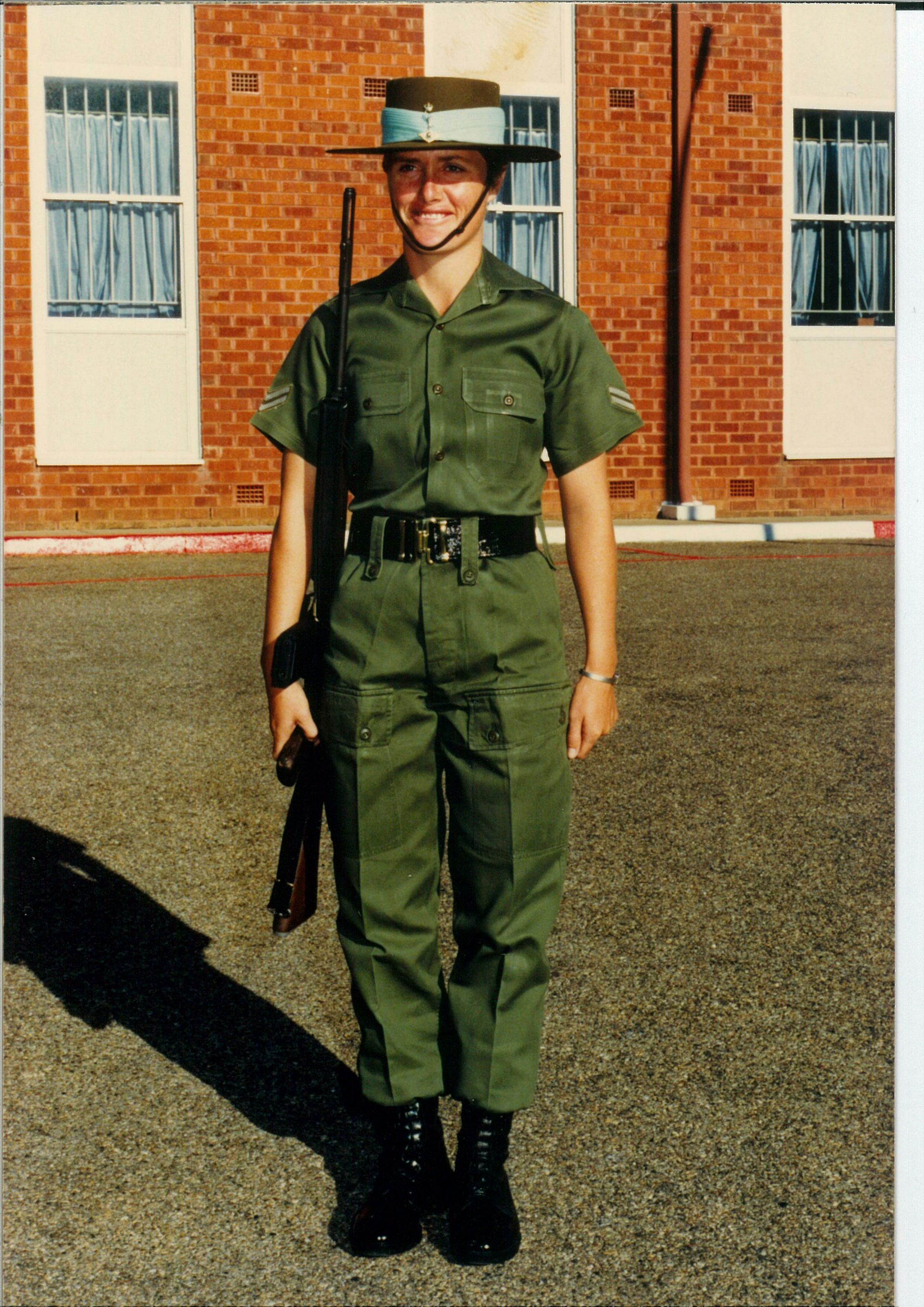 A young woman in Army uniform and a hat with a blue band, holding a rifle, standing to attention and smiling.