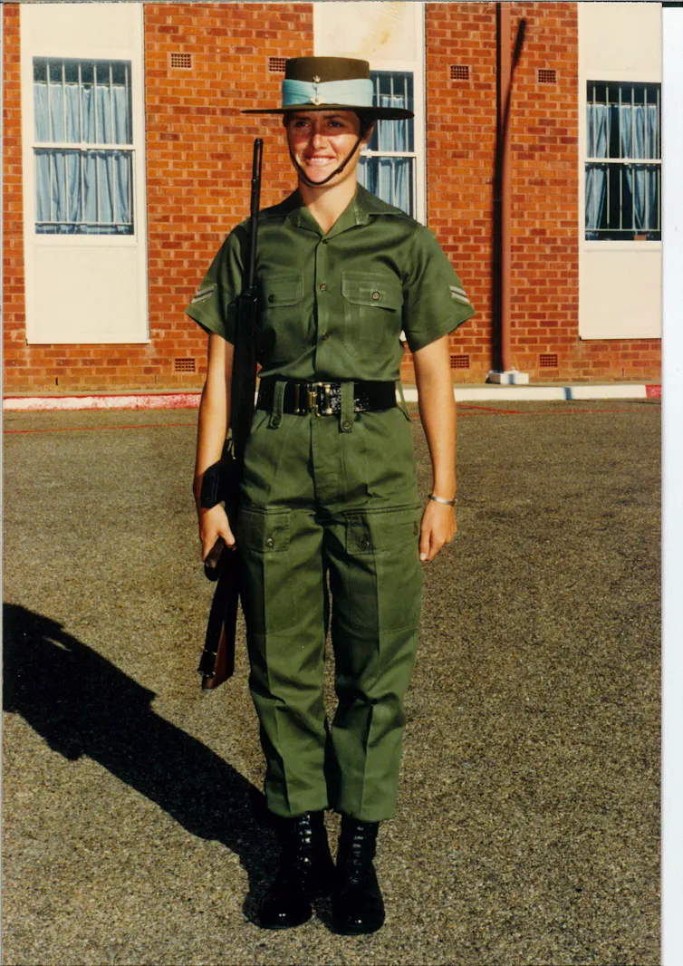 A young woman in Army uniform and a hat with a blue band, holding a rifle, standing to attention and smiling.
