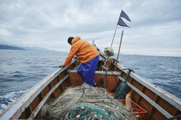 A man leans on the side of a boat filled with fishing nets, on the ocean